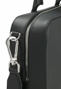 Sac en cuir noir avec une finition texturée, fermeture éclair argentée et attache à clip en métal. Le design est angulaire et compact.