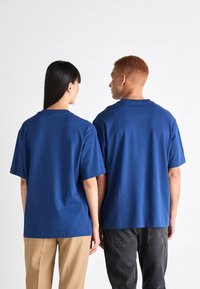 Camisetas de algodón azules de manga corta, de corte relajado, con costuras visibles y cuello redondo, llevadas por dos personas que miran hacia el lado opuesto de la cámara.