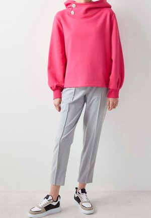Roze katoenen hoodie met wijde mouwen en een hoge kraag met twee witte knopen. Gecombineerd met grijze op maat gemaakte broek en zwart-witte sneakers.