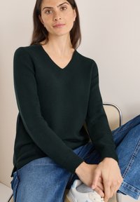 Dunkelgrüner V-Ausschnitt-Pullover mit langen Ärmeln, aus weichem Material. Kombiniert mit blauen Jeans und weißen Turnschuhen. Einfaches Design und Textur.