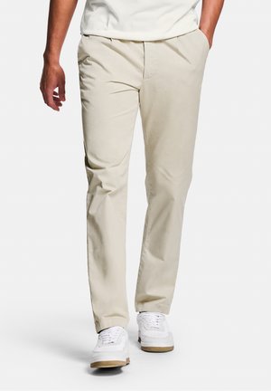 Mann geht, trägt beigefarbene Chinos und weiße Sneakers, mit einer Hand in der Tasche und einem teilweise sichtbaren weißen Hemd.