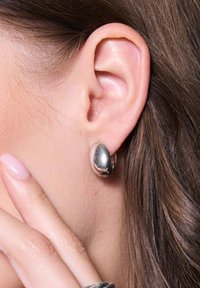 Srebrna metalowa kolczyk w formie gładkiej, zaokrąglonej bryły. Umieszczony przy uchu, podkreślający jego wypolerowaną powierzchnię i odbicie.