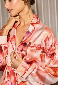 Cămașă de pijama din mătase florală cu un fundal roz, având flori mari roșii și purpurii, margini zimțate și un buzunar pe piept cu logo brodat.