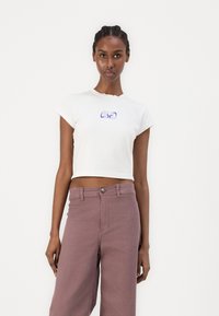 White cropped t-shirt with a scalloped neckline, featuring blue script "OBEY" logo, paired with light purple high-waisted pants.