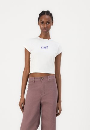 Hvid cropped t-shirt med en bølget udskæring og med blå skrift "OBEY" logo, parret med lys lilla højtaljede bukser.