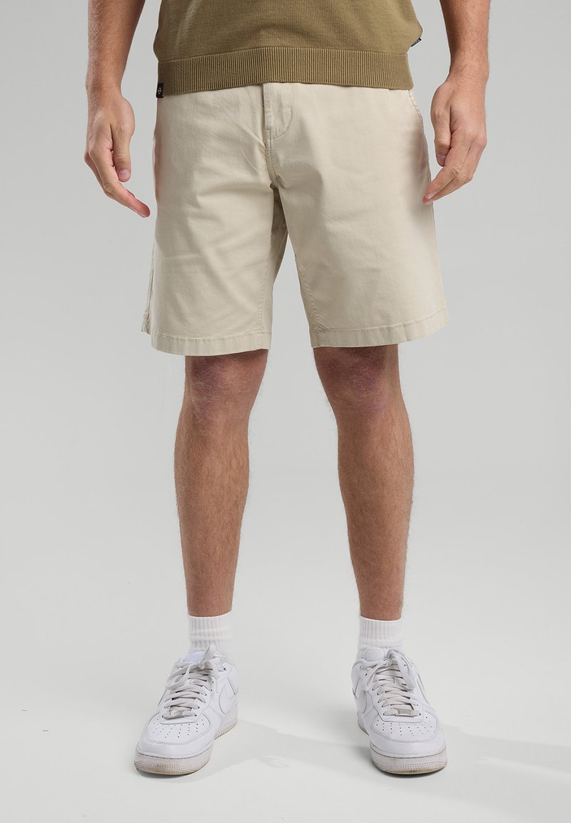 Beige cotton shorts with a straight cut, side pockets, and double-stitched hem. Paired with white sneakers and ribbed green sweater.
