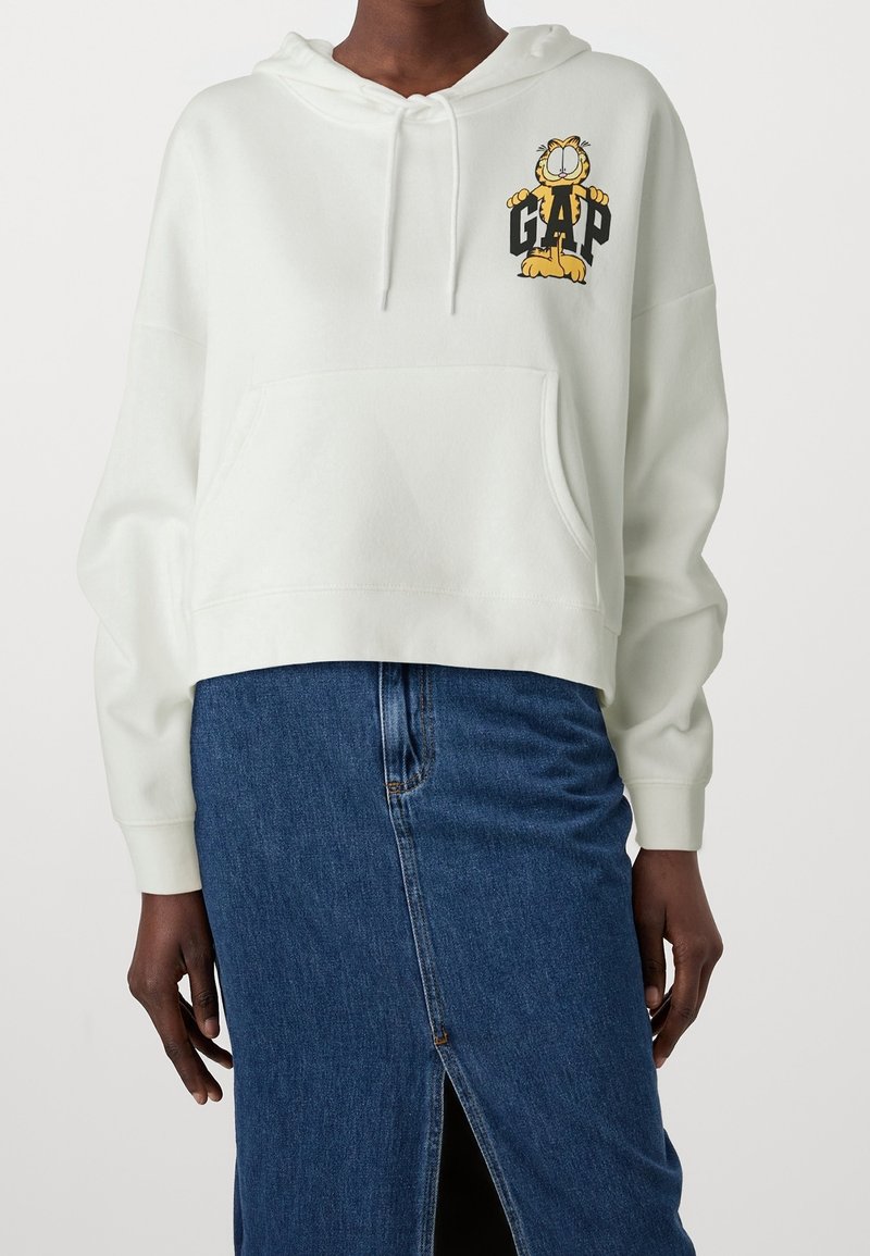 Corta sudadera blanca con bolsillo frontal y un gráfico de un personaje de dibujos animados. Material ligero, mangas largas y capucha con cordón.