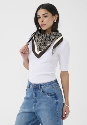 Žena s dlhými tmavými vlasmi, oblečená v krátkemu bielom tričku s krátkym rukávom, modrých džínsoch a čierno-bielom šále so vzorom, drží okraj šálu.