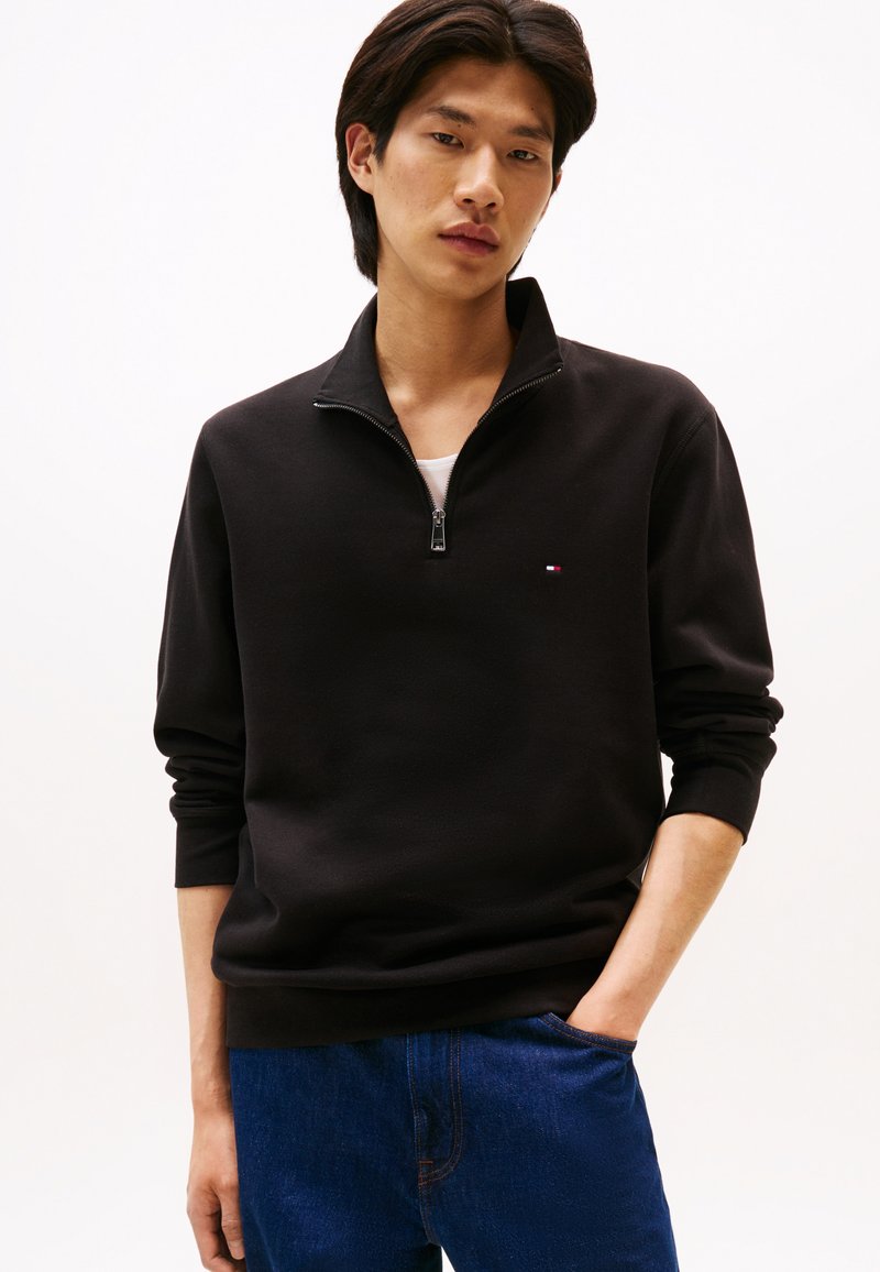 Ung mand iført en sort sweatshirt med kvart lynlås og blå jeans, med den ene hånd i lommen mod en ensfarvet lys baggrund.