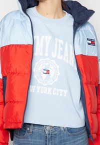 Persona vestita con una giacca a vento rossa e azzurro chiaro sopra un maglione azzurro chiaro di Tommy Jeans e jeans di denim blu.