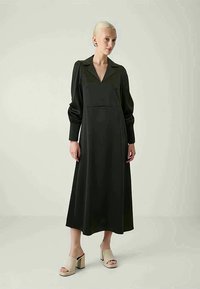 Vestido longo preto com colarinho, mangas longas bufantes e corpete ajustado. Usado com sandálias de plataforma bege. Textura suave e design clássico.