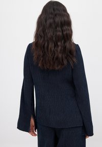 Chaqueta texturizada azul marino con mangas largas de campana, que presenta rayas verticales y aberturas en los costados del dobladillo para mayor movilidad.