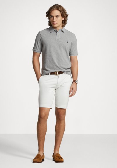 Pilkas polo marškinėliai trumpomis rankovėmis, balti šortai, rudas odinis diržas ir šviesūs loaferiai. Modelis stovi su rankomis kišenėse.