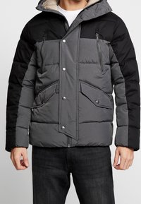 Giacca imbottita in grigio e nero. Presenta una tasca sul petto con cerniera, chiusura frontale con bottoni automatici e grandi tasche laterali. Interno morbido.