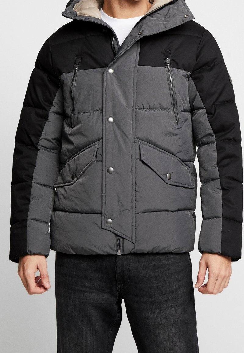 Giacca imbottita in grigio e nero. Presenta una tasca sul petto con cerniera, chiusura frontale con bottoni automatici e grandi tasche laterali. Interno morbido.