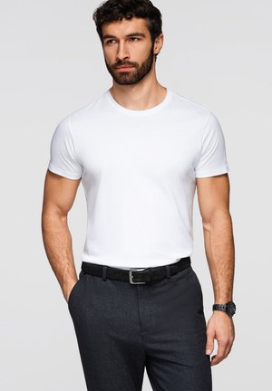 Viiksimies, joka pukeutunut yksinkertaiseen valkoiseen lyhythihaiseen t-paitaan, tummaan vyöhön, tummiin tekstuuriin koristeltuihin housuihin ja mustaan rannekelloon, seisoo yhdellä kädellä taskussa.