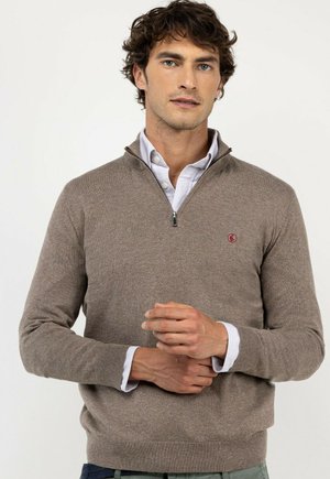 Hombre con un suéter marrón claro con cremallera hasta el pecho sobre una camisa blanca con cuello, con las manos entrelazadas al frente sobre un fondo liso.