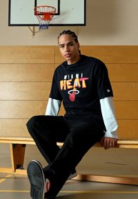 Camiseta negra con el logo "MIAMI HEAT" en colores brillantes, superpuesta sobre una camiseta blanca de manga larga. Pantalones negros y zapatillas negras con acentos rojos.
