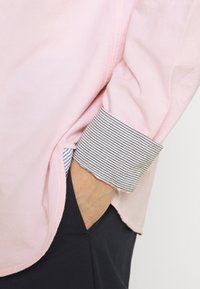 Camicia rosa chiaro con dettaglio di polsino a righe, tessuto morbido, vestibilità comoda, spacco laterale, abbinata a pantaloni scuri.