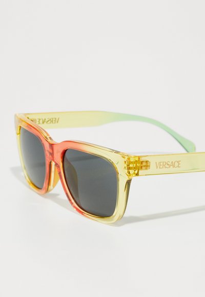 Versace saulės akiniai su permatomu geltonu ir raudonu rėmu, tamsiais objektyvais ir žaliai tonuotais šoneliais; ant šventokos yra logotipas.