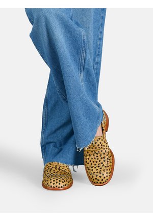 Scarpe slip-on con stampa leopardata in un colore giallo-marrone con macchie nere, abbinate a pantaloni in denim blu a gamba larga con orlo sfrangiato.