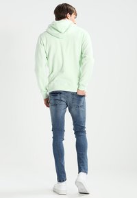 Mintgrüner Kapuzenpullover mit Kordelzug, kombiniert mit engen blauen Jeans und weißen Sneakers, zeigt einen lässigen, entspannten Look.