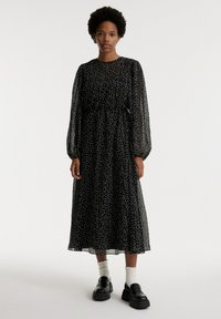 Zwart langemouwen jurk met witte stippen, doorschijnende stof, verzamelde taille en enkel-lange zoom. Gecombineerd met chunky zwarte schoenen en witte sokken.