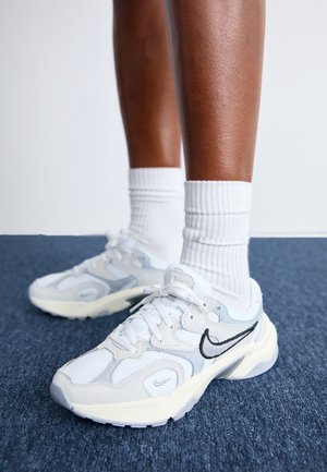 Persoon draagt witte en grijze Nike sneakers met zwart Swoosh-logo en witte geribbelde crew-sokken op een blauw tapijt.