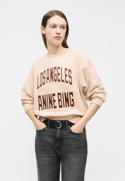 Kvinne iført beige genser med teksten «Los Angeles Anine Bing», svart belte og mørkegrå jeans, hendene i lommene, ser litt til siden.