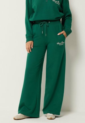 Persona che indossa pantaloni della tuta verdi a gamba larga e felpa abbinata con scarpe da ginnastica bianche, una mano in tasca, in piedi davanti a uno sfondo semplice.