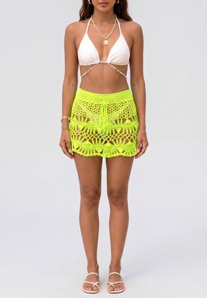 Femme portant un haut de bikini blanc, une mini-jupe jaune néon en crochet, des sandales blanches, un collier en or et des bracelets en or, debout devant un fond uni.