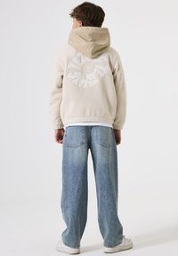 Chaqueta de forro polar beige con detalle del logo blanco en la parte posterior, llevada sobre una camisa blanca, combinada con jeans anchos de color azul claro y zapatillas blancas.