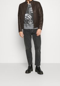 Giacca di pelle marrone con colletto alto, dettagli a spalla testurizzati, abbinata a una maglietta grafica, jeans grigi e stivaletti neri alla caviglia.