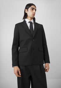 Black suit jacket with a tailored fit, single button closure, two front pockets, and a smooth texture. Worn with a white shirt and black tie.