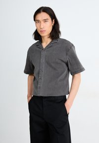 Short-sleeve, gray button-up shirt in a faded denim fabric with a notched collar and front pocket detail, paired with black pants.
