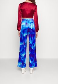 Žena v živých modrých vzorovaných širokých kalhotách, v krátkém červeném saténovém topu a v bílých lodičkách na kotníku, stojící na jednobarevném pozadí.