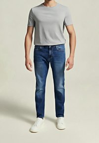 Harmaa lyhythihainen t-paita, siniset farkut haalistuneilla yksityiskohdilla ja valkoiset slip-on-kengät, joita esittelee seisova henkilö neutraalia taustaa vasten.