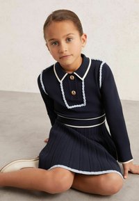 Marineblaues Strickkleid mit langen Ärmeln, weißem Kragen und geflochtenen Akzenten. Plissierter Rock und zwei Knöpfe an der Brust. Glatte, gestrickte Textur.