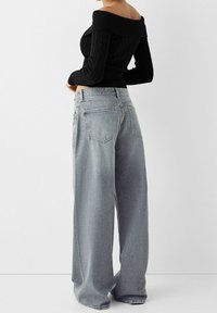 Kvinna klädd i en svart off-shoulder kabelstickad topp och ljusblå högmidjade, vida jeans, stående mot en neutral bakgrund.