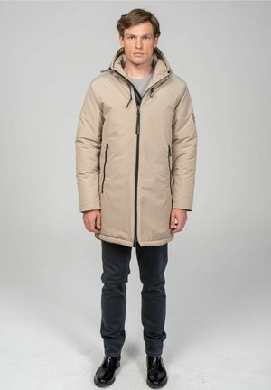 Beige padded coat with a zip front, adjustable hood, and two side pockets. Worn over a gray shirt and paired with dark pants and shiny black shoes.