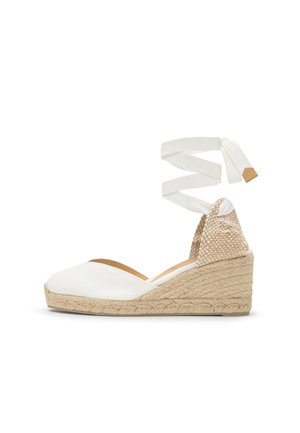 Sleehak espadrille met een beige gevlochten hak, witte gesloten neus en lange witte enkelwikkelbanden op een witte achtergrond.