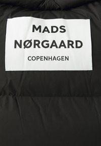 Doudoune noire avec une étiquette blanche sur laquelle est inscrit en noir "MADS NØRGAARD COPENHAGUE." Texture lisse avec un détail de logo.