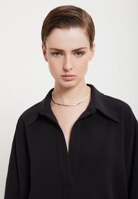 Musta kauluspaita syvällä V-päällä, jossa on sileä kangas ja huomaamaton ompelu. Yhdistetty hopeakolorikaulaketju, jossa on kuusikulmaisia yksityiskohtia.