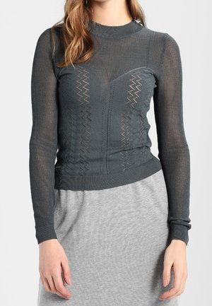 Vrouw draagt een donkergrijs, langarmig, semi-doorzichtig gebreid shirt met zigzagpatronen, gecombineerd met een lichtgrijze rok.