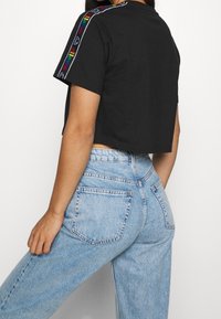 Camiseta negra corta con acentos de estampado colorido a lo largo de los hombros, combinada con jeans de cintura alta azul claro con bolsillos traseros.