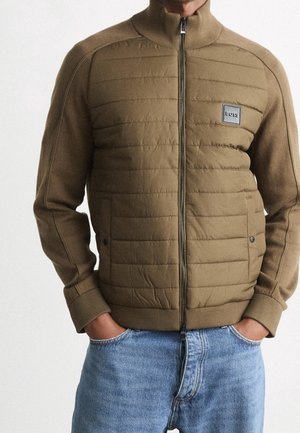 Mann trägt eine braune gesteppte Jacke mit Reißverschluss, Rippbündchen und Kragen, steht mit den Händen in den Taschen der blauen Jeans.