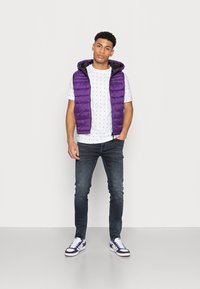 Paarse gewatteerde vest met capuchon over een witte T-shirt met een klein maritiem patroon, gecombineerd met donkere spijkerbroeken en witte sneakers.