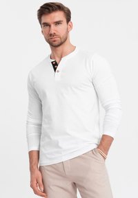 Valkoinen pitkähihaisen t-paita, jossa on musta nappikaulus, jossa on kolme ruskeaa napia. Sileä kangas, slim fit -malli, yhdistetty vaaleisiin housuihin.