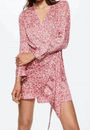 Kvinde iført en pink blomsterbindekjole med lange ærmer og V-hals, stående med den ene arm krydset over kroppen.