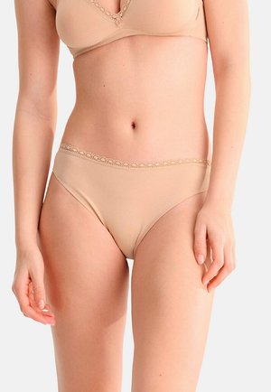 Femme portant un soutien-gorge beige orné de dentelle et des sous-vêtements assortis, debout devant un fond neutre, les bras détendus le long du corps.